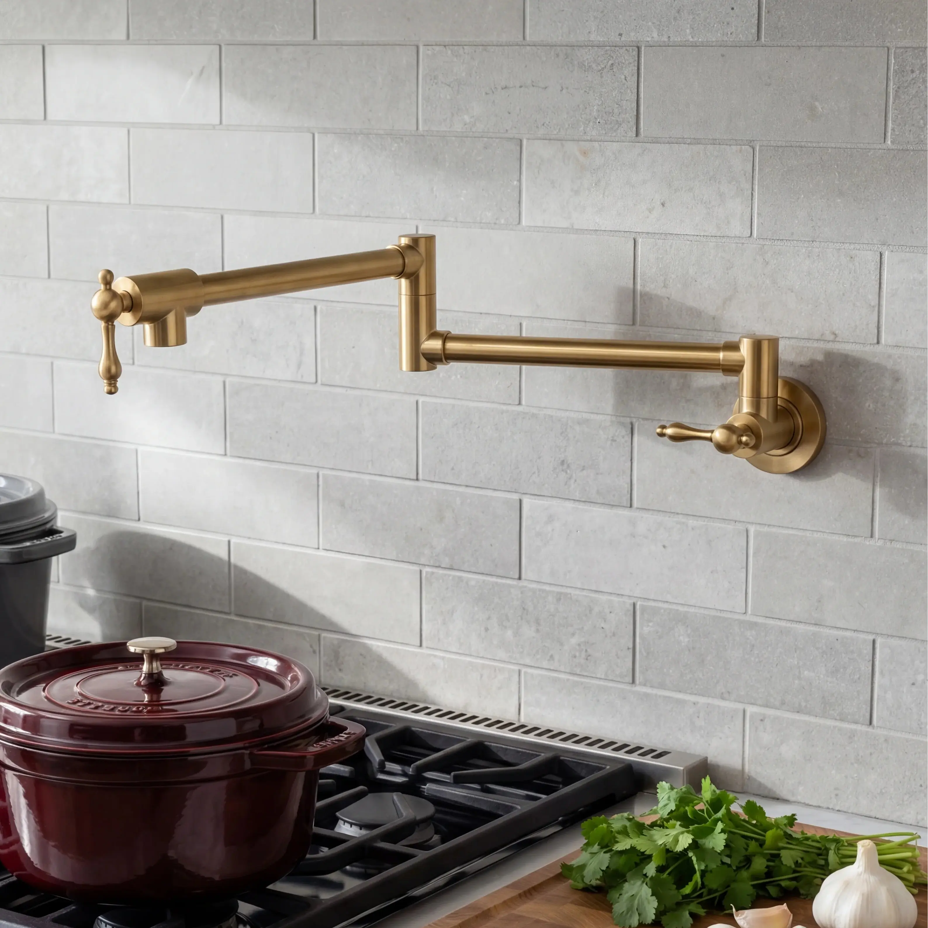 A wall-mounted brass kitchen faucet with an adjustable gooseneck spout and separate lever handle, installed on a light gray subway tile backsplash above a gas stove. A dark red enameled pot sits on the burner, with fresh parsley and garlic on the counter.